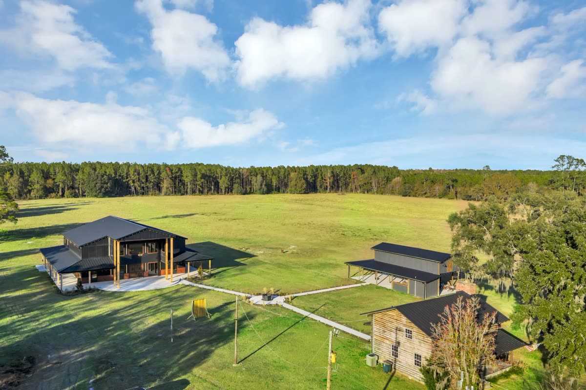An aerial view of the ranch: The Venue, the Pole Barn and the log cabin set in open pasture against a pine treeline