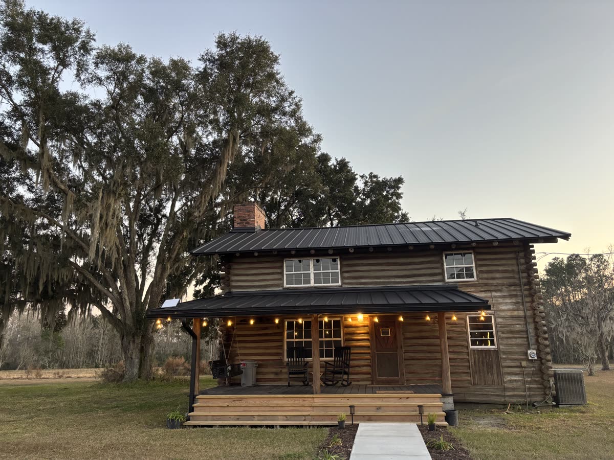 The log cabin at dusk beneath a Spanish moss oak, porch lights glowing