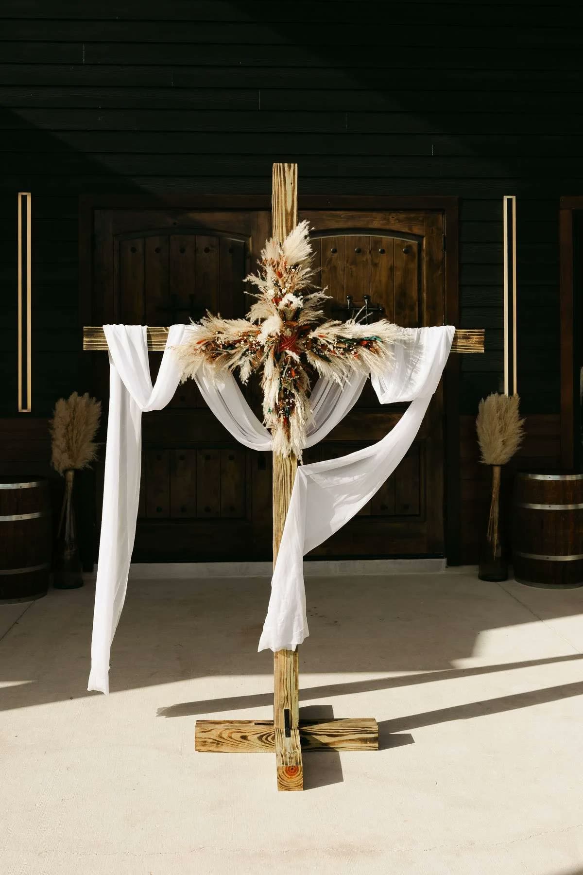 A wooden cross draped in white fabric and pampas grass against the hall's dark entry doors
