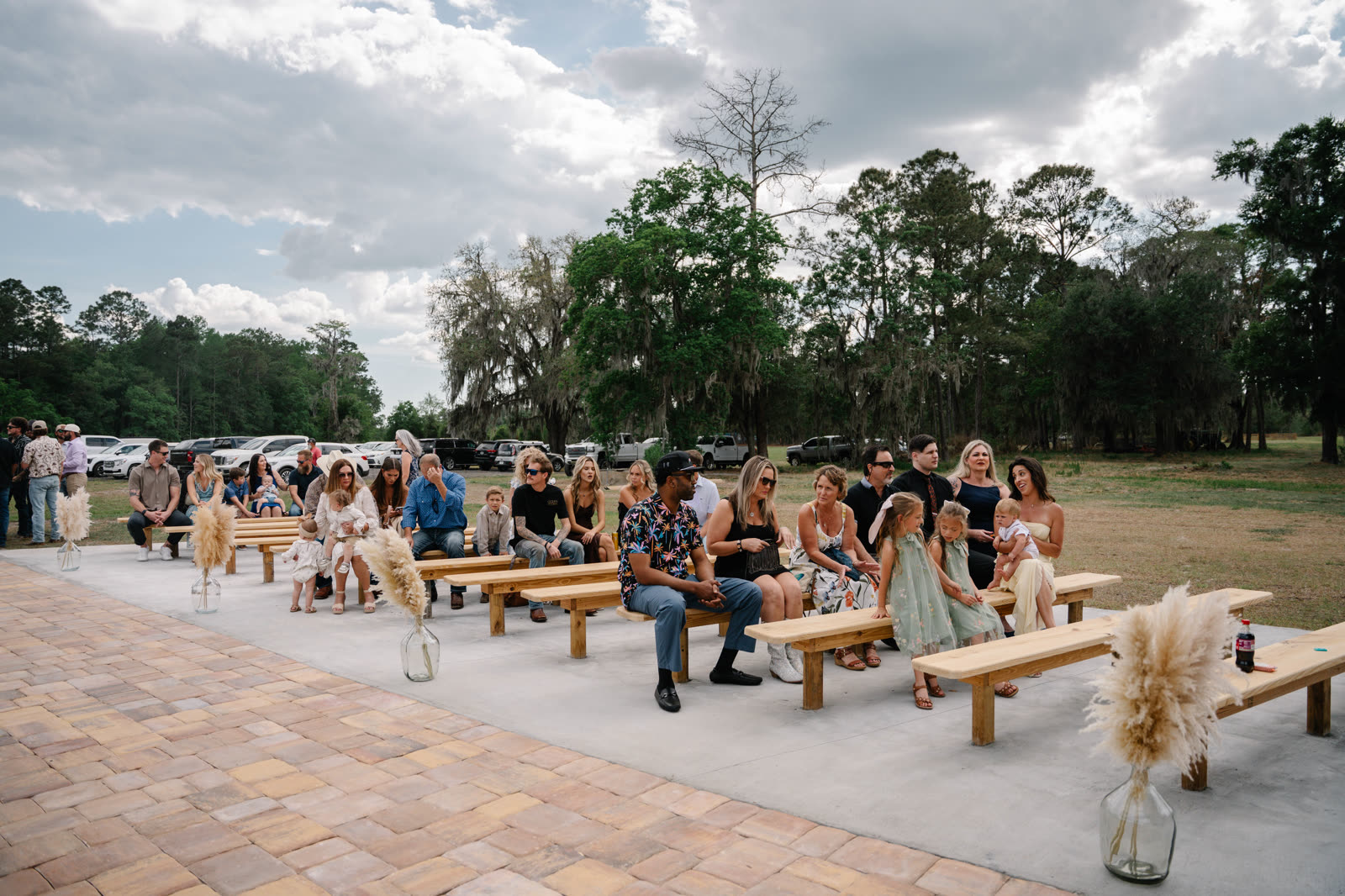 Guests seated on wooden benches along the paver aisle for an outdoor ceremony under the oaks