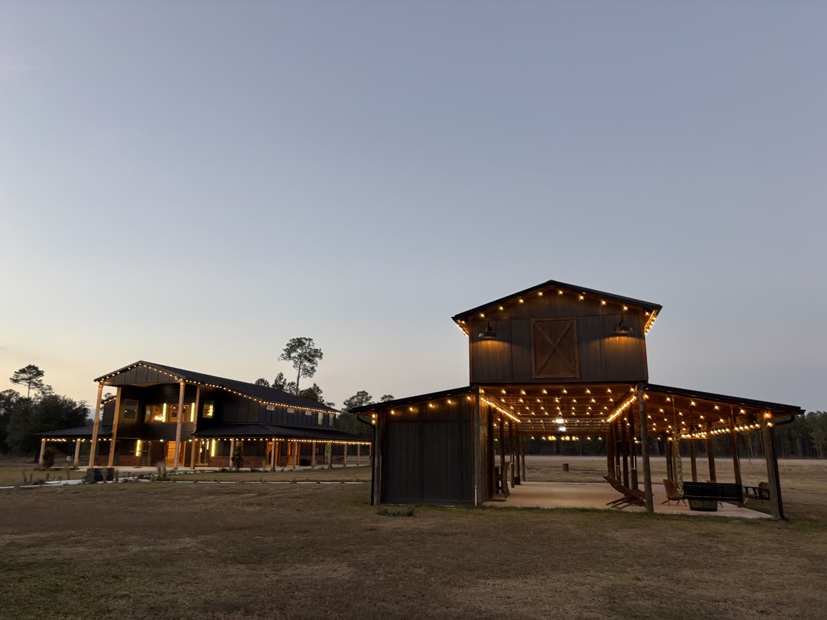 The Pole Barn at dusk, its gable and open bays outlined in warm string lights across the pasture