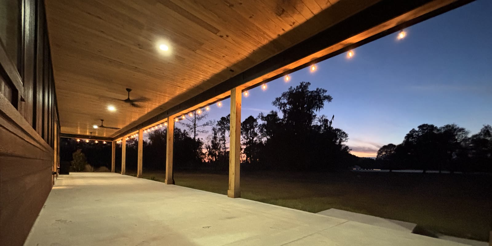 The covered veranda at Three Fours Ranch at dusk, string lights running its length toward a pink sunset over the pasture