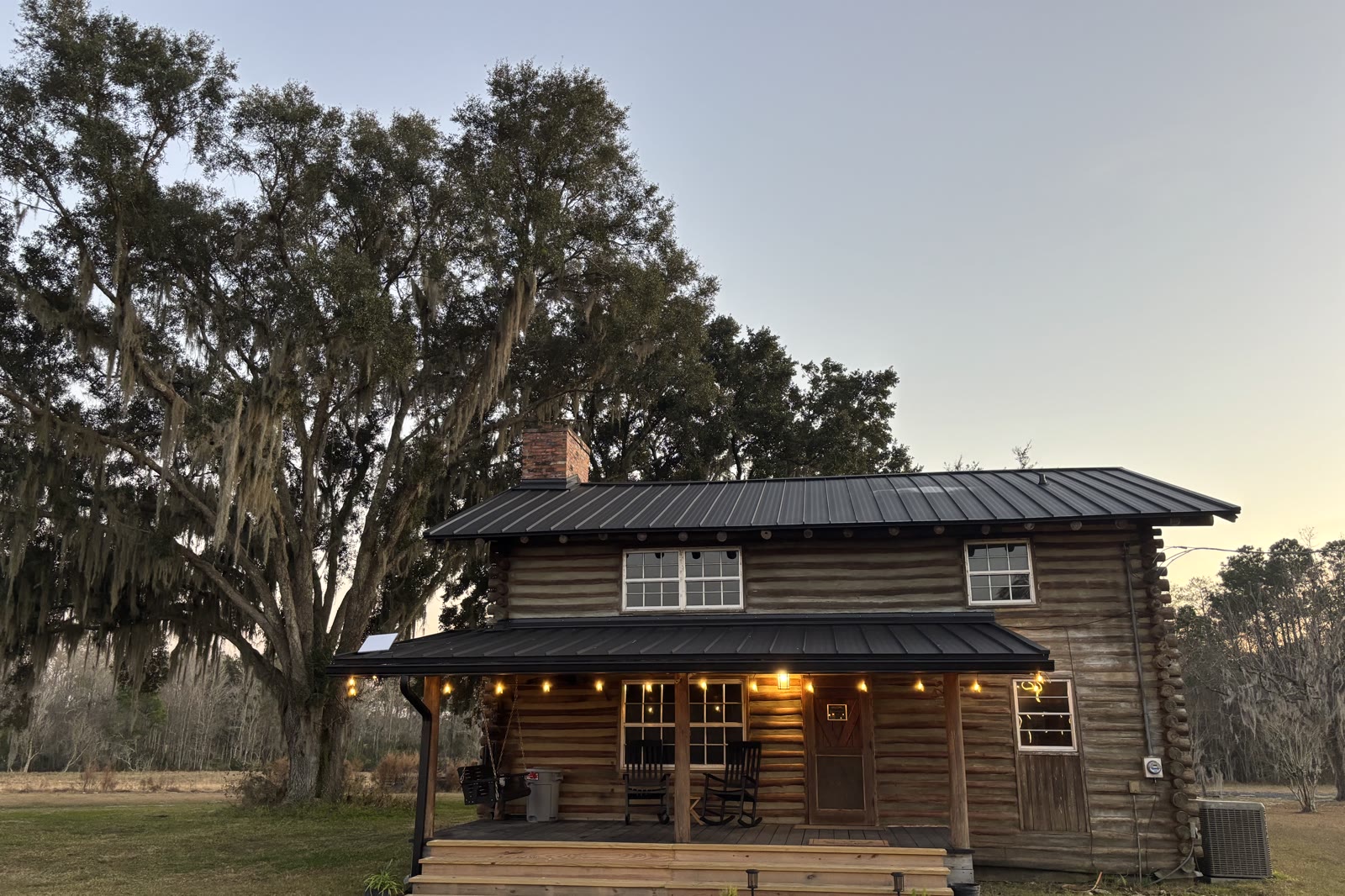 The log cabin at Three Fours Ranch at dusk, porch lights glowing beneath a Spanish moss oak