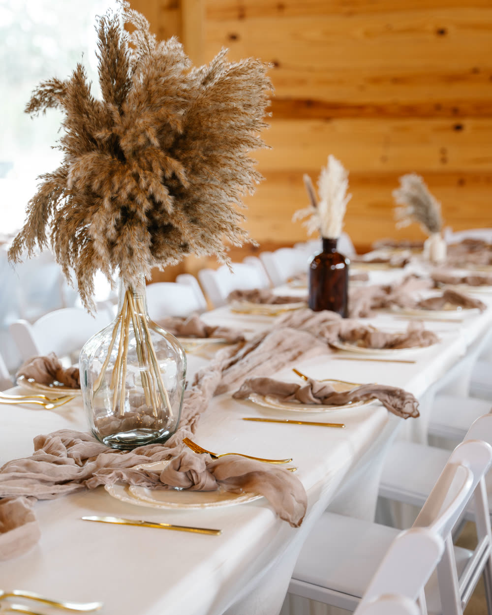A long banquet table set with gold flatware, blush gauze runners and pampas grass